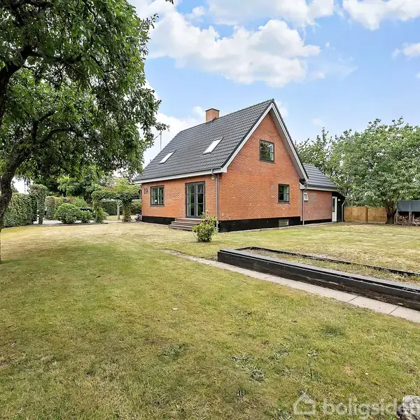 Rødt hus med sort tag står i en velholdt have. Træer omkranser haven, og der er en grussti forrest i billedet. Skydfri himmel giver et klart lys.