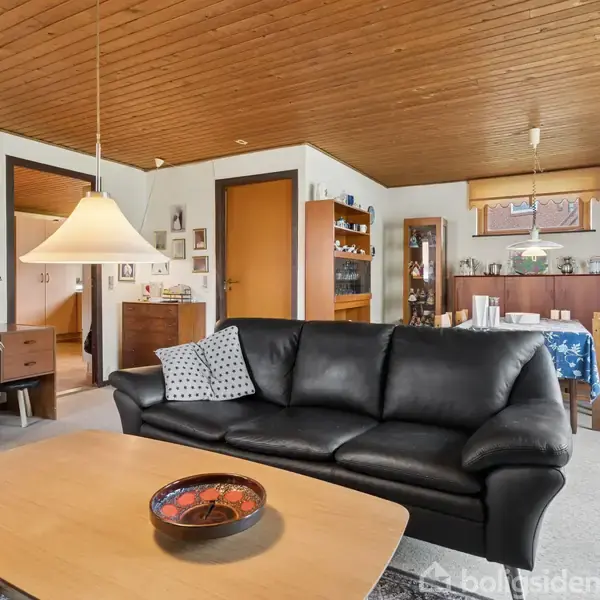 Sort lædersofa står i en stue med trægulv og loft, omringet af skrivebord, malerier på væggen, lys træhylder og et spisebord med dug.