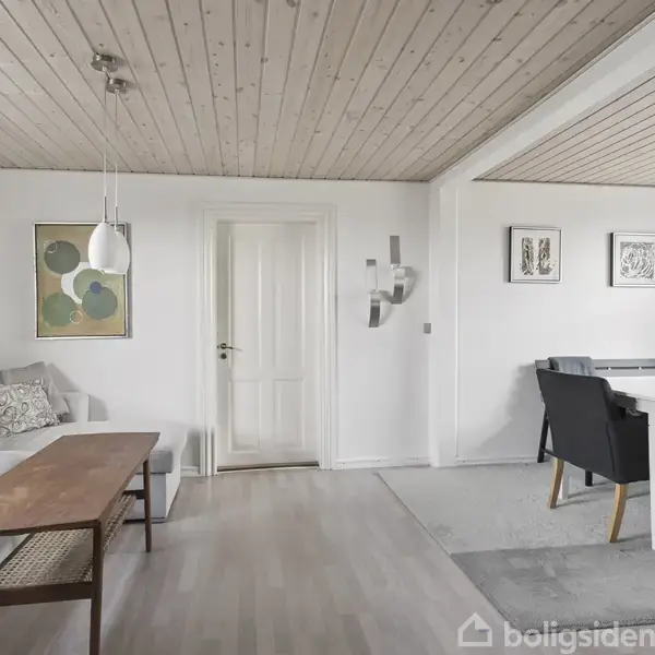 Stue med grå sofa og træbord, hængende lamper, og malerier på hvide vægge. Ved siden af er et spiseområde med bord og sorte stole, omgivet af lyst trægulv og loft.