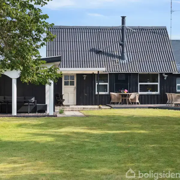 Et sort hus med en terrasse, hvor to borde og stole står. En græsplæne og en overdækket terrasse er i forgrunden, omgivet af træer og buske.