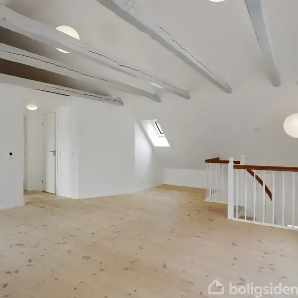 Et lyst loftsrum med lys trægulv. Loftsbjælkerne er synlige, og der hænger en rund lampe. En trappe fører nedad, og et ovenlysvindue ses til højre.