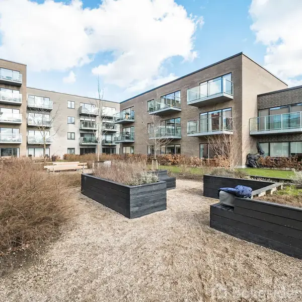 Lejlighedsbygning omkranser en gårdhave med højbede og grusbelagt gangsti; har flere altaner og moderne arkitektur. Skydækket himmel i baggrunden, omgivet af beplantning.
