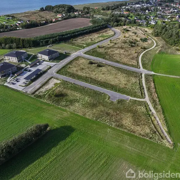 Nogle bygninger står omkring nyanlagte veje ved et udviklingsprojekt, omringet af grønne marker og tæt på en by. En sø kan ses i baggrunden.