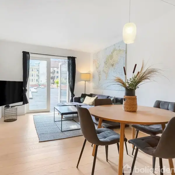 Rundt spisebord med stole står i en lys stue med trægulv. I baggrunden ses en sofa, vægkort og en standerlampe ved altandøren, der har sorte gardiner.