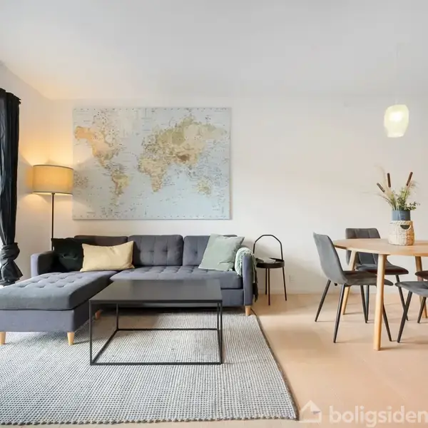 Gråt sofaarrangement står ved siden af et spisebord i moderne stue; kort over verden hænger på væggen; store vinduer til venstre giver udsigt til terrasse udenfor.