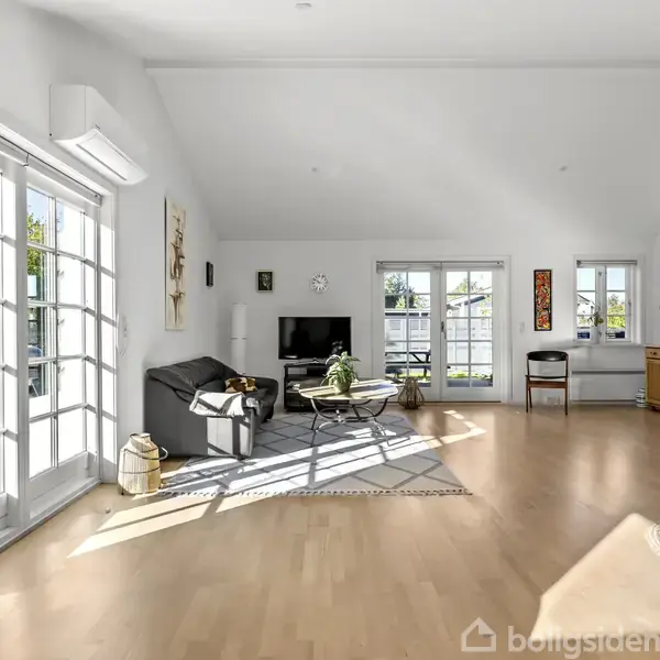 En lys og rummelig stue har en sort sofa og tv i et hjørne nær glasdøre, der åbner til udsigt. En kommode står ved vinduet med blomsterdekorationer. Trægulv skaber en varm atmosfære.
