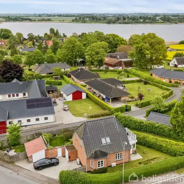 Flere huse ligger langs en vej i en grøn forstad. En stor sø er i baggrunden med marker og træer omkring.