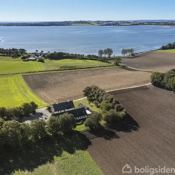 En stor landejendom omgivet af marker ligger tæt ved en sø. En lang allé træer fører op til bygningen. I baggrunden ses en vid udsigt over sø og landskab.