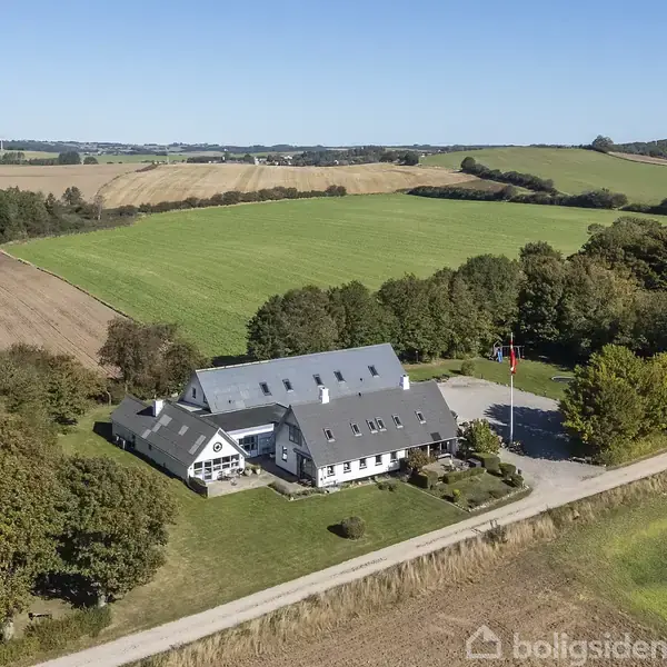 En stor gård med hvide bygninger og grå tage står omgivet af marker og træer. Bygningen har en stor gårdsplads med flagstang. Landskabet er bakket og landligt.