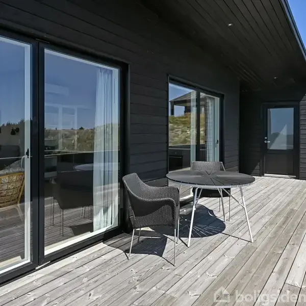 Terrassemøbler bestående af et rundt bord og to stole står på en træterrasse. I baggrunden er der store glasdøre og et sort træhus med blå himmel over.
