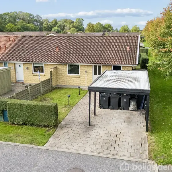 Rækkehus med rødt tag står ved siden af en carport med skraldespande. En grøn hæk omkranser plænen, og til højre er en vej med rækker af træer.