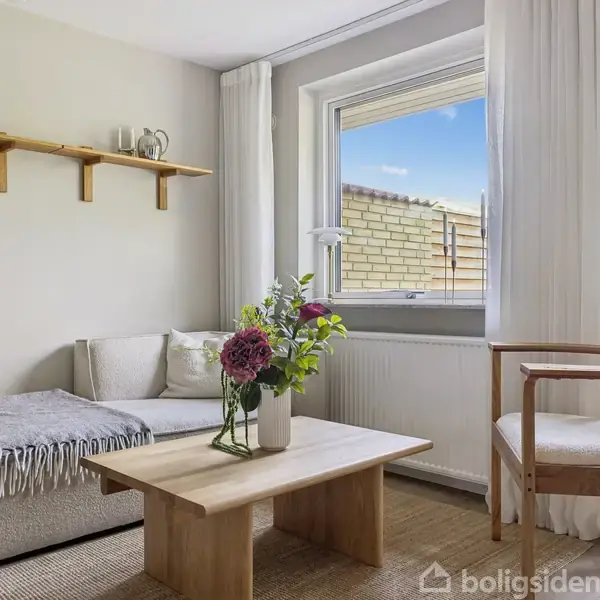 Stue med en lysegrå sofa og træbord, blomster i vase på bordet, hylde med dekorationer på væggen, brede vinduer med udsigt til have, indrettet med neutrale farver.
