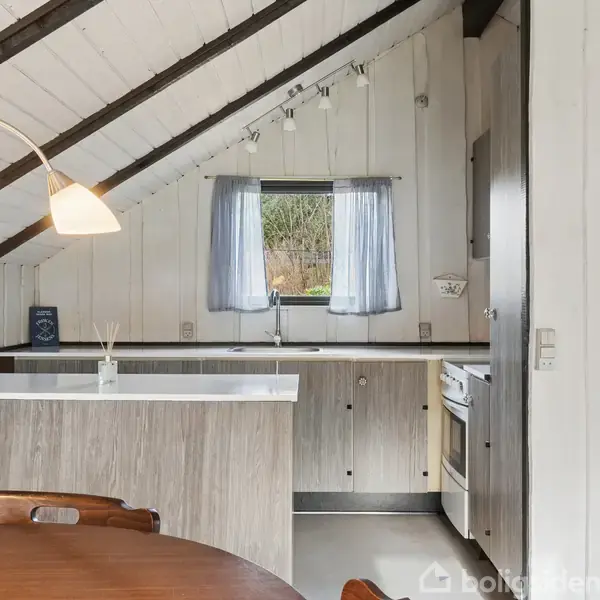 Et køkken med træbeklædte skråvægge og loft. Bordpladen er hvid, og der er et lille vindue med gardiner. Rundt spisebord med blomster i forgrunden.