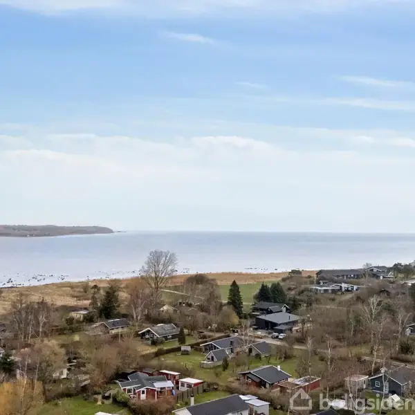 Kystnære huse ligger spredt blandt træer og grønne områder, mens de grænser op til en bred strandlinje ved havet under en klar blå himmel.