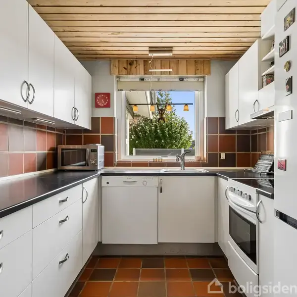 Et moderne køkken med hvide skabe, sort bordplade og brune fliser. En hvid ovn står ved siden af et køleskab med magneter. Et vindue viser grønt haveområde udenfor.