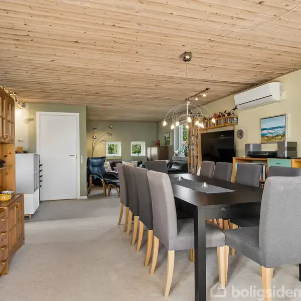 Et stort, sort spisebord med grå stole står i et rum med trægulv og loft. Flere træreoler fyldt med forskellige genstande fylder væggen, og der er en aircondition.