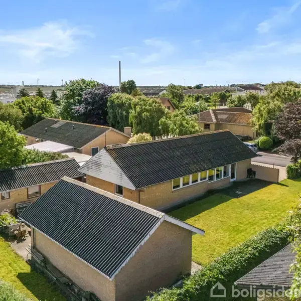 En enkeltfamiliehus med mørkt tag ligger i et grønt boligområde. Huset har en stor have med græsplæne og omkringliggende hække. Andre huse ses i baggrunden.