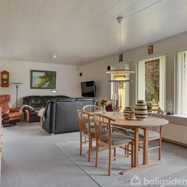 Stue med spisebord, stole og sofaer. Rummet har store vinduer, malerier på væggene og en gammel standur. Lyseblå vægge og tæpper skaber en hyggelig atmosfære.