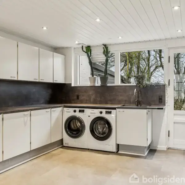 Vaskemaskine og tørretumbler står under en bordplade i et lyst vaskerum med hvide skabe. Vinduet og døren til haven giver udsigt til træer udenfor.