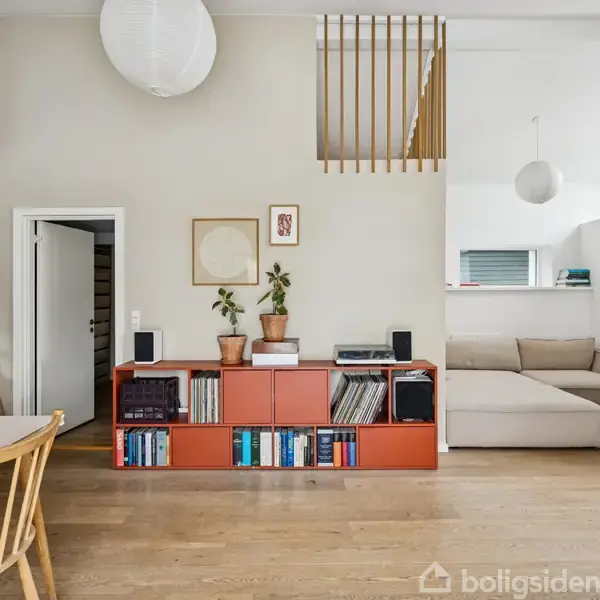 Reol med planter og bøger står foran en lys væg ved siden af en stige. Til højre er en sofa med krumformet design og en brændende pejs i et moderne opholdsrum.