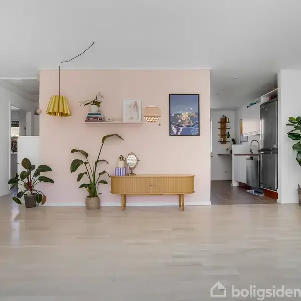 Stue med lyserød accentvæg. Hylde og maleri på væggen; gulvlampe og planter tilføjer dekoration. Moderne køkken ses i baggrunden, åben og minimalistisk indretning.