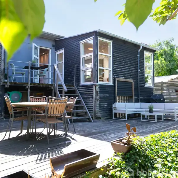 Sort træhus med terrasse har store vinduer i kontrast til lysegrå møbler og spisebord. En trappe fører til en hævet balkon. Planter tilføjer grønne omgivelser.