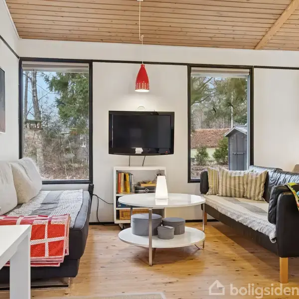 Stue med to sofaer omkring et rundt bord; en fladskærm hænger på væggen. Rummet er lyst med store vinduer, der viser en have udenfor.