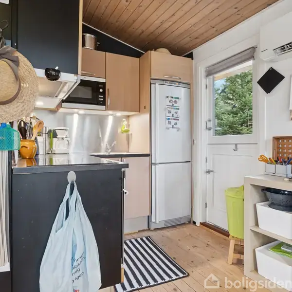 Køkken med komfur og køleskab, omgivet af lyse træmøbler og en grøn skammel ved vinduet. En stråhat hænger ved siden af en hylde med forskellige kogegrej.