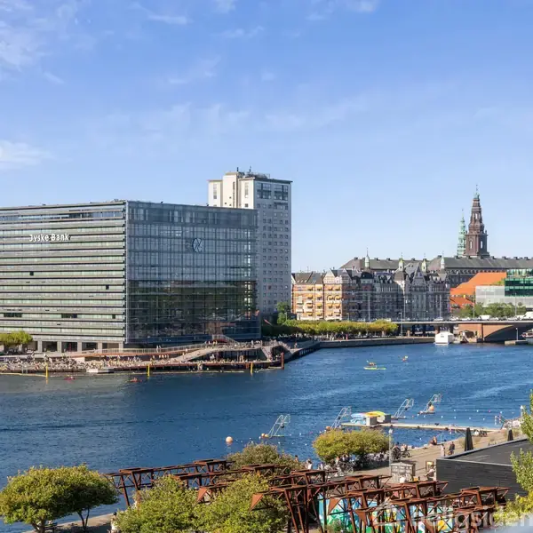 Et stort glasbyggeri står ved siden af en bred flod, hvor der er små både med mennesker. I baggrunden ses en by med spidse tårne under en blå himmel.
