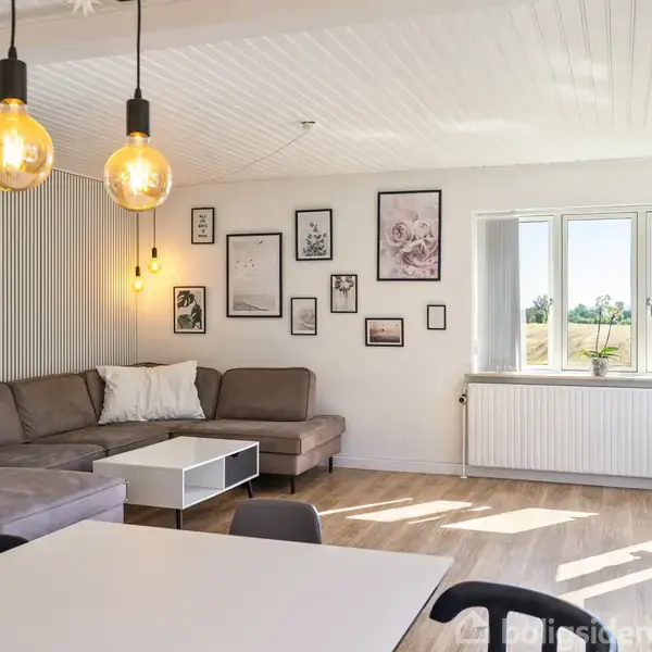 En stue med en grå hjørnesofa er oplyst af hængende glødepærer. Billeder hænger på væggen, og store vinduer viser udsigt til et grønt landskab.