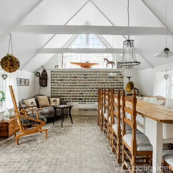 Et stort, lyst rum med loft til kip, indeholder en lang træspisebord med stole. I baggrunden ses en sofagruppe og et spejl. Væggene har hylder og dekorative elementer.