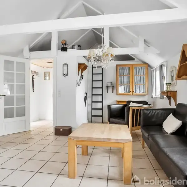 Stue med sort lædersofa og træbord, flisebelagt gulv og loft med synlige bjælker. Hylder og vinduer på væggene. En gynge stol står ved den hvide dør.