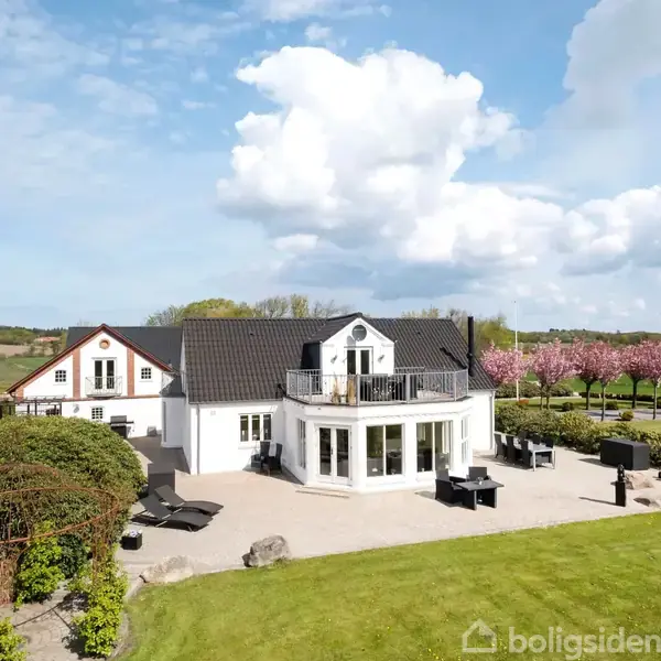 Et hvidt hus med sort tag står midt i en velplejet have, omgivet af grønne områder og blomstrende træer under en blå himmel med skyer.