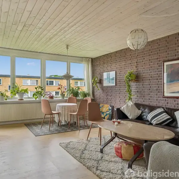 Stue med sofa ved murstensvæg, sofabord, og tæppe; spisebord med fire stole ved vindue med udsigt til gule bygninger udenfor. Grønne planter og kunst pryder rummet.