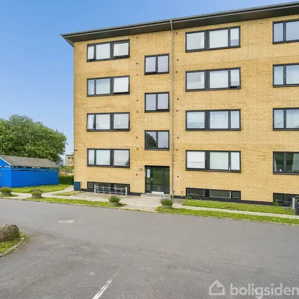 Et gult murstenshøjhus med sorte vinduesrammer står stille i et boligområde, omgivet af grønne plæner. En blå bygning ses til venstre, og der er cykler parkeret ved siden.