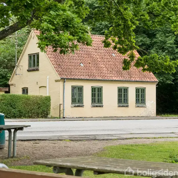 Et gult murstenshus med rødt tegltag står ved siden af en vej. Huset har grønne vinduer og er omgivet af træer og buske.