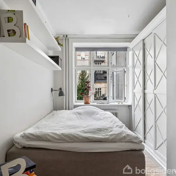 Et lille soveværelse med en seng dækket af en hvid dyne, placeret op ad en væg. Der er hylder med bøger og en lampe over sengen. Et åbent vindue med en plante udenfor. En garderobe står langs den anden væg.