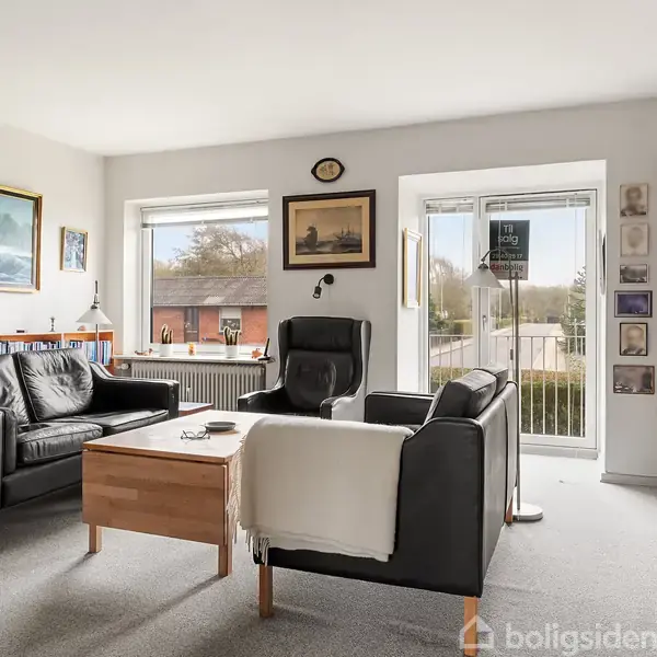 Et hyggeligt opholdsområde med sorte lædersofaer og sofabord, omgivet af reoler fyldt med bøger. Store vinduer bader rummet i naturligt lys, og vægkunst dekorerer væggene.