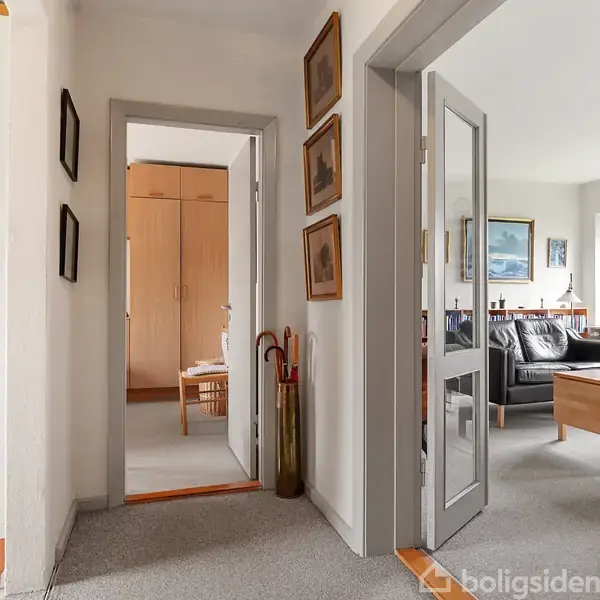En gang med tæppebelagt gulv fører til flere rum. Døre står åbne til en stue med sofa og billeder på væggene samt et andet rum med træskabe. Paraplyer står ved siden af dørkarmen.