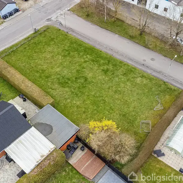 Åbent græsareal, omgivet af hække, bag et hus med sort tag. En trampolin og en gynge er placeret i haven. Vej og bygninger ses i baggrunden.