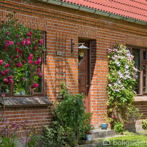 Murstenshus med rødt tegltag og klatreroser langs facaden. En trælaterne hænger ved hoveddøren. Haven har frodigt grønt og blomsterbed.