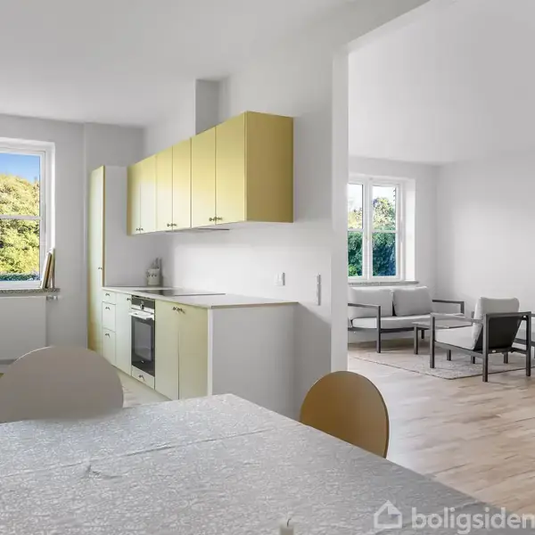 Et køkken-alrum med et spisebord i forgrunden og gulv-til-loft vinduer, der giver udsigt til grønne omgivelser. Køkkenet har lysegule skabe og en ovn i hjørnet. En stue med sofaer ses i baggrunden.