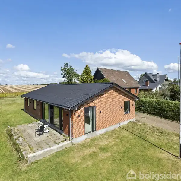 Rødt murstenshus med sort tag står stille på en grøn græsplæne med terrasse, omkranset af hække. Landskabet omkring inkluderer marker, en flagstang og flere huse i baggrunden.