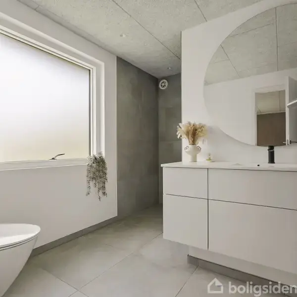 Badeværelse med hvidt møbel, rund spejl over vask og planter som dekoration. Store vindue giver naturligt lys. Toilettet er monteret på væggen. Rummets design er minimalistisk.
