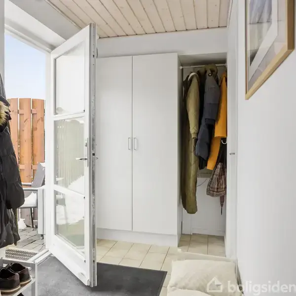 En dør til venstre står åben mod en gårdhave med trægulv. Skabe fyldt med jakker i lys entré. Sko på gulvet og en pude liggende ved siden af døren.