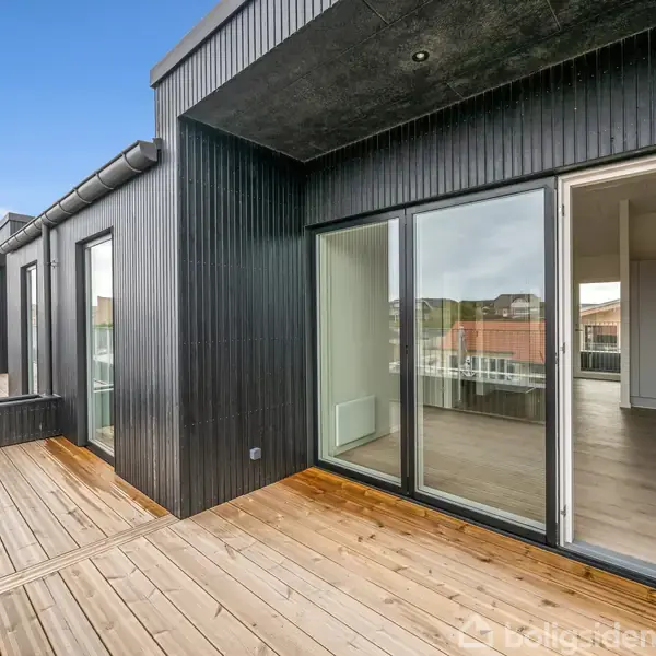 Træterrasse foran moderne, sortmalet hus med store glasdøre, der åbner mod et lyst værelse. Bygninger og himmel i baggrunden. Ingen tekst i billedet.