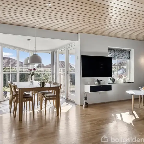 Et moderne stueområde med et spisebord og stole står ved glasvægge med udsigt til en have. En sofa og sofabord er i højre side, en fladskærm hænger på væggen.