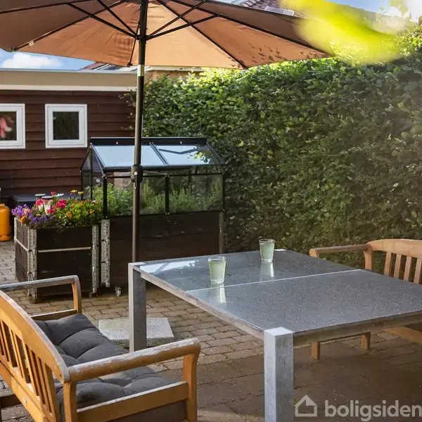 Borde og stole står under en stor parasol på en terrasse. I baggrunden er en grill, blomsterkasser, et mini-drivhus, og en hæk, der skaber en hyggelig haveatmosfære.