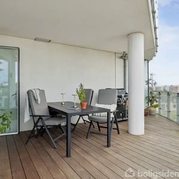 En stor, moderne altan med trægulv har et sort bord med stole arrangeret omkring det. Et lille træ og planter er placeret i nærheden. Baggrunden viser byudsigt og glasrækværk.