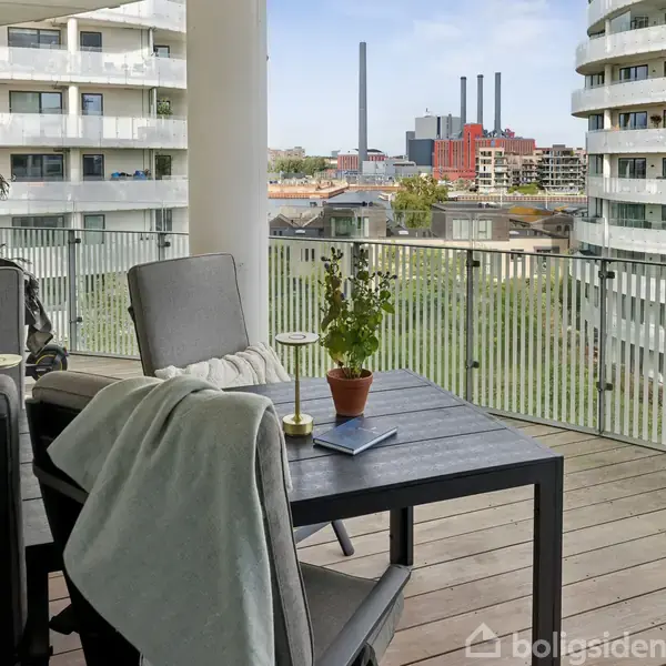 Altan med spisebord og stole dækket af et tæppe, omgivet af moderne lejlighedskomplekser. En plante i en potte står på bordet. Baggrunden viser byens panorama.